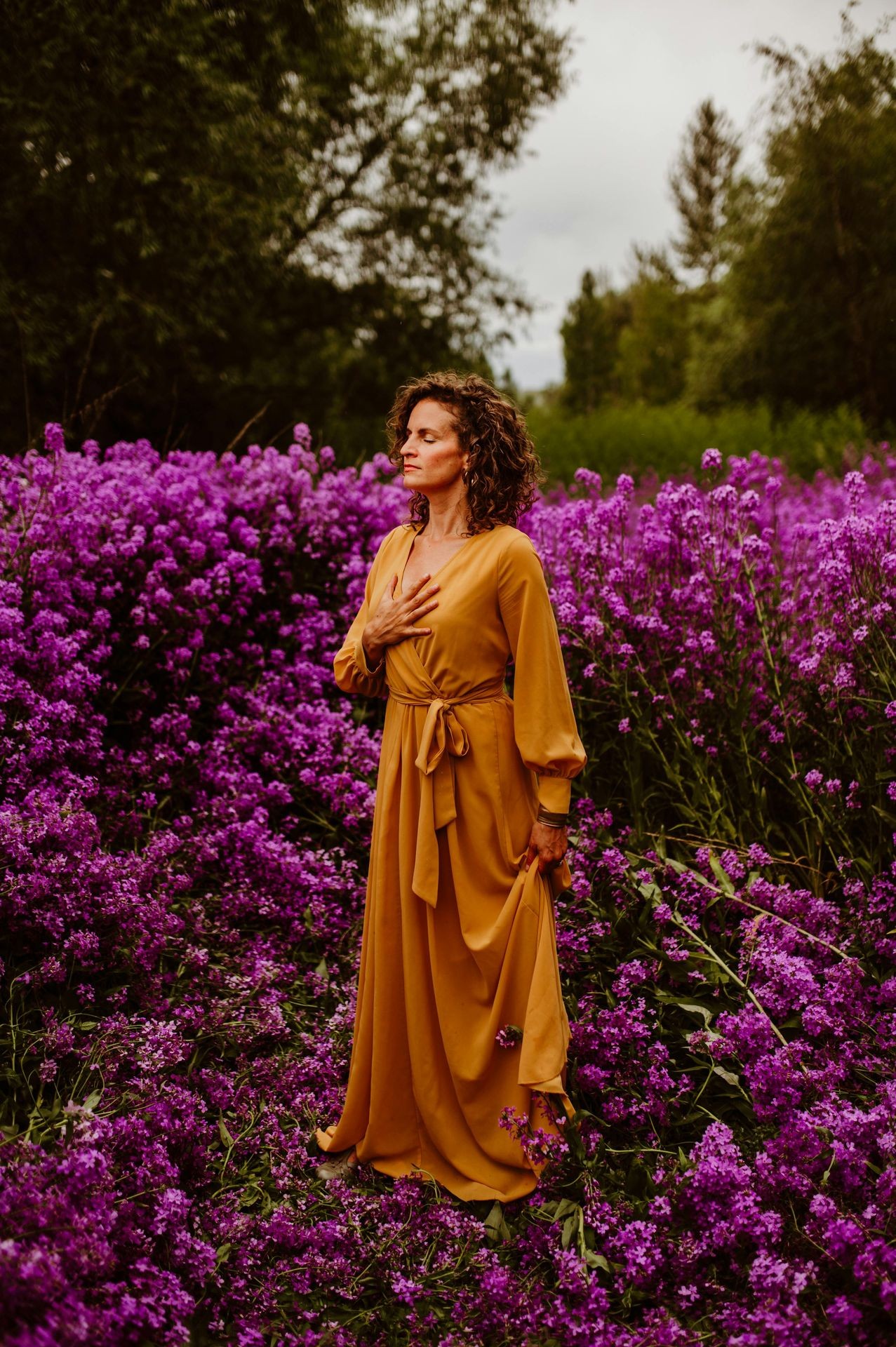 Woman in yellow dress stands among vibrant purple flowers in a lush garden.