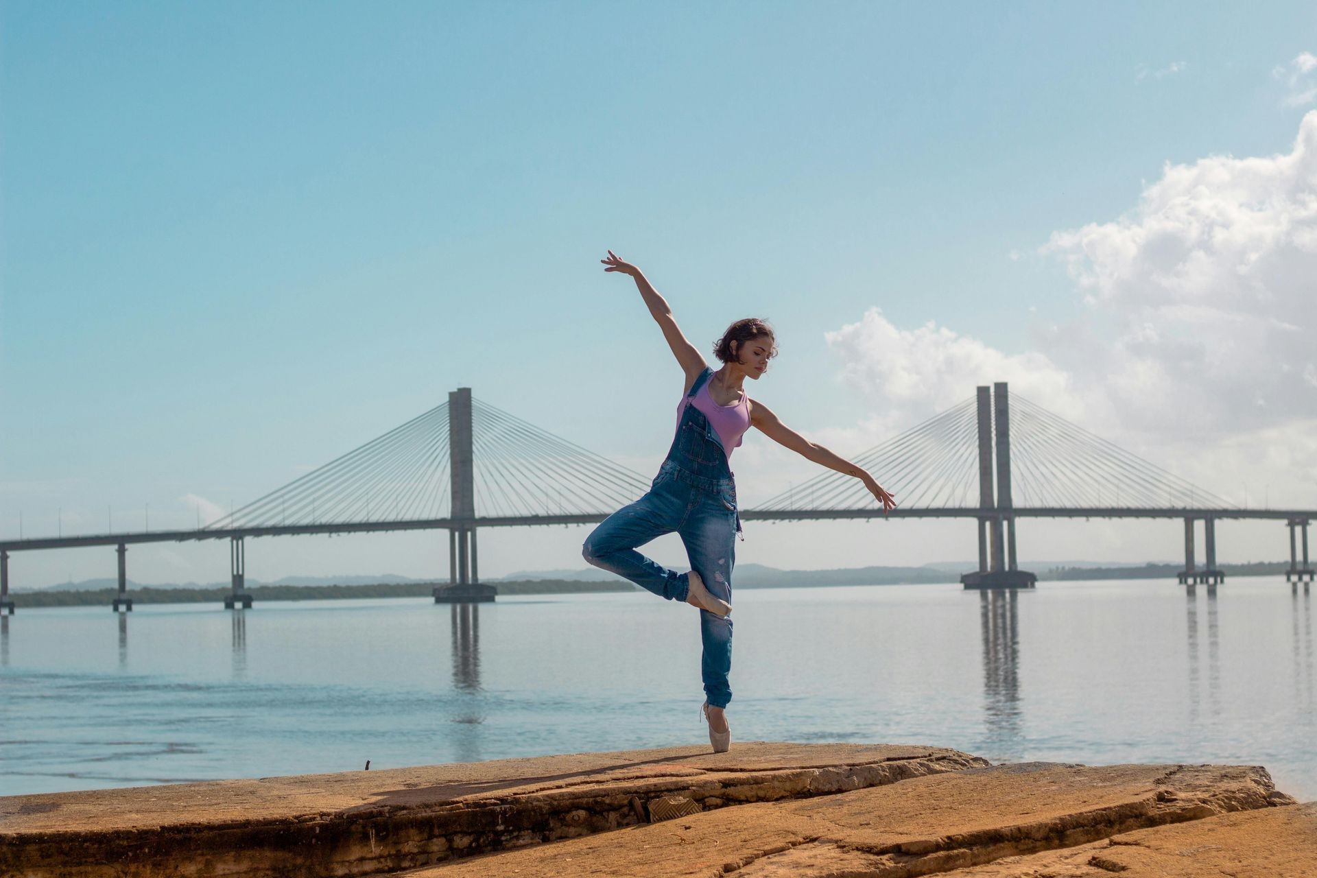 Person dancing gracefully on rocks near a large bridge over water, with a clear blue sky.